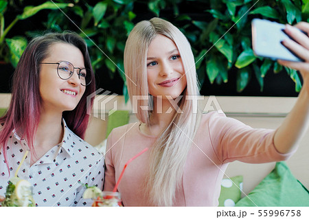 Two young beautiful girls, a blonde in a pink dress and a brunette with dyed hair, sit at a cafe Two young beautiful girls, a blonde in a pink dress and a brunette with dyed hair, sit at a cafe 55996758