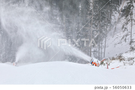 Orange and blue snow making cannon spreading ice crystals over ski piste, trees in background 56050633