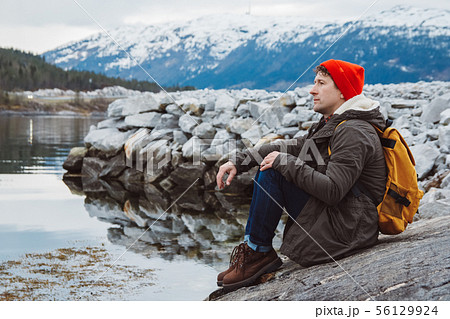 Traveler man with a yellow backpack wearing a red hat sitting on the shore on the background of 56129924