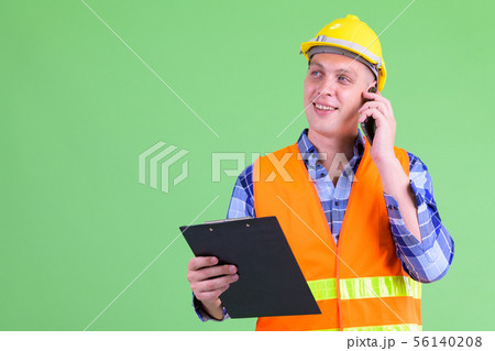Happy young man construction worker talking on the phone while holding clipboard Happy young man construction worker talking on the phone while holding clipboard 56140208