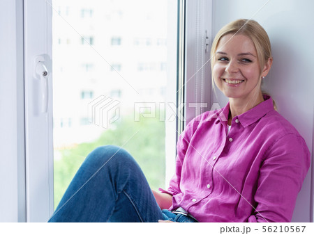 Pretty blonde girl sitting on a window and smartphone in hands Pretty blonde girl sitting on a window and smartphone in hands 56210567