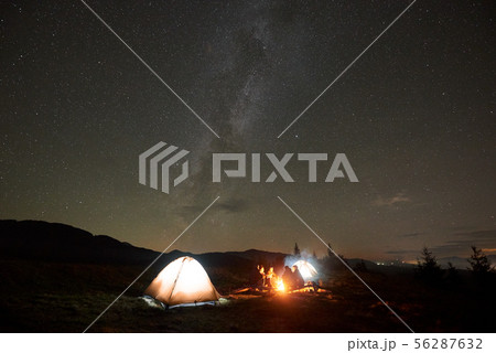 Group of tourists with guitar by burning bonfire under dark starry sky with Milky Way constellation. 56287632