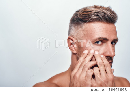 Skin problems. Worried man with a stubble examining his face and looking at camera while standing Skin problems. Worried man with a stubble examining his face and looking at camera while standing 56461898