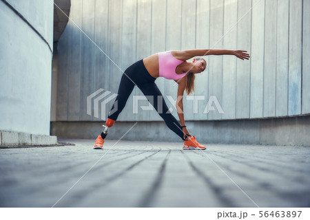 Great shape. Young woman with leg prosthesis in sportswear stretching before morning workout while 56463897