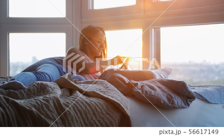 Young cheerful woman using smartphone on the bed during sunset by the window 56617149