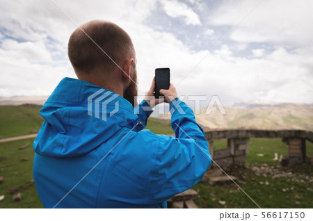 A bearded man takes photos or videos outdoors on his smartphone. A guy in a jacket on a background A bearded man takes photos or videos outdoors on his smartphone. A guy in a jacket on a background 56617150