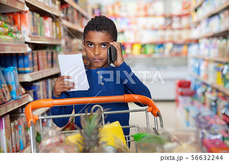 Focused African tween boy talking on phone and looking at shopping list while visiting supermarket Focused African tween boy talking on phone and looking at shopping list while visiting supermarket 56632244