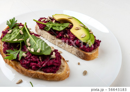 Bruschettas with beetroot hummus, avocado, spinach and seeds on a white plate on a white background. Bruschettas with beetroot hummus, avocado, spinach and seeds on a white plate on a white background. 56643183