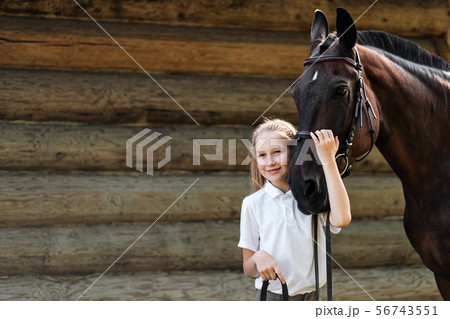 A teenage girl jockey stands next to a brown horse and hugs her. Against the background of a wooden 56743551