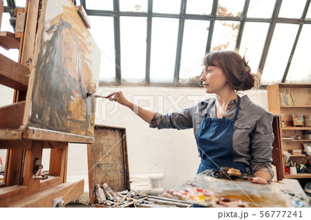 Woman artist paints a picture on canvas. Bright art studio with a large window. Easels and canvases. 56777241