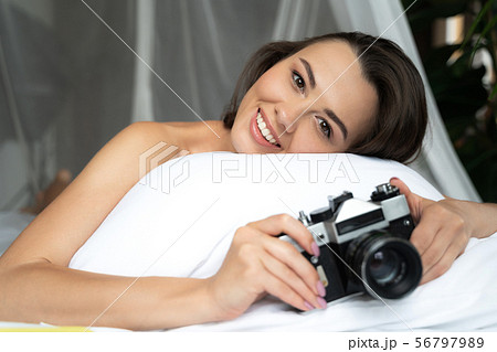Joyful young woman lying in bed and holding retro camera Joyful young woman lying in bed and holding retro camera 56797989