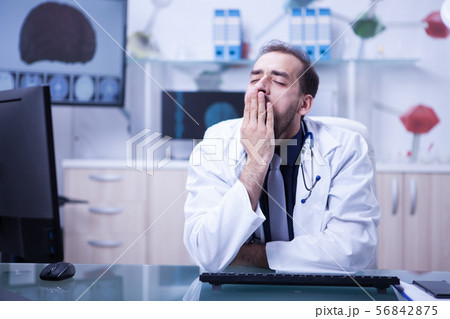 Young bearded doctor tired and yawning sitting at the office in his hospital cabinet 56842875