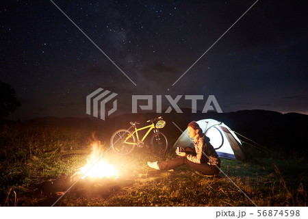 Woman resting at night camping near campfire, tourist tent, bicycle under evening sky full of stars 56874598
