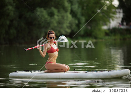 Young pretty fitness girl in swimwear is paddling on a SUP in the middle of the lake with a green Young pretty fitness girl in swimwear is paddling on a SUP in the middle of the lake with a green 56887038
