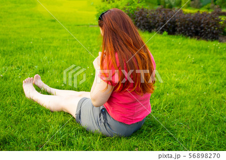 Red-haired girl sits with his back on the grass in the park. Vacation and vacation concept. Red-haired girl sits with his back on the grass in the park. Vacation and vacation concept. 56898270