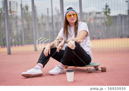Image of athletic brunette girl in sunglasses sitting on skateboard on playground on summer day 56920930