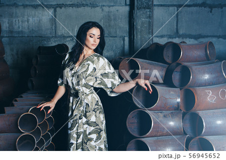 model business concept, heavy industry, building materials, beautiful brunette with bright red manicure, in dress and shoes posing among metal pipes. A woman in beautiful clothes, standing in the ware model business concept, heavy industry, building materials, beautiful brunette with bright red manicure, in dress and shoes posing among metal pipes. A woman in beautiful clothes, standing in the ware 56945652