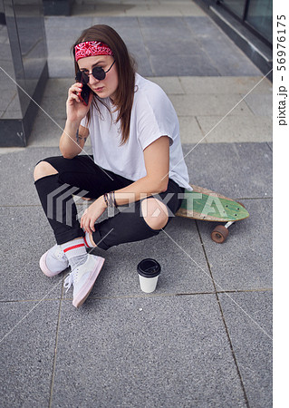 Photo of young brunette in sunglasses talking on phone sitting on skateboard on background of modern Photo of young brunette in sunglasses talking on phone sitting on skateboard on background of modern 56976175