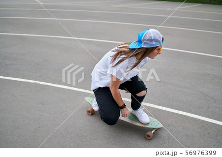 Photo on side of young brunette woman in baseball cap riding skateboard outdoors 56976399