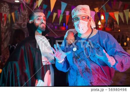 Scary man in a doctor costume covered in blood with his handsome dracula friend celebrating 56980498