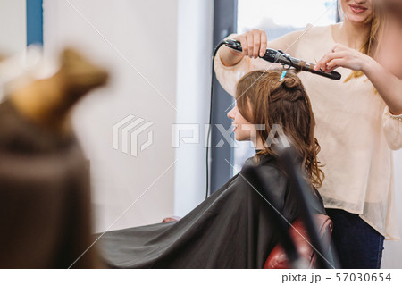 beauty, hairstyle concept, happy young woman and hairdresser with hair iron making hairdo at hair 57030654