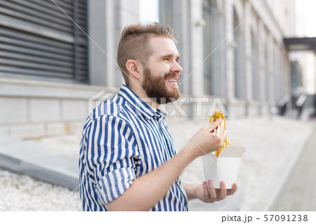 Close-up of an amusing young hipster guy eating chinese noodles with wooden chopsticks sitting in a Close-up of an amusing young hipster guy eating chinese noodles with wooden chopsticks sitting in a 57091238