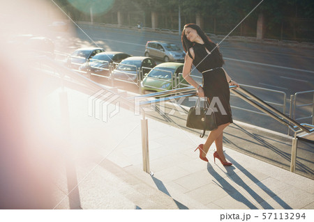 beautiful brunette in a black dress with a bag walking down the street in the light. Woman advertises accessories, cosmetics, makeup 57113294