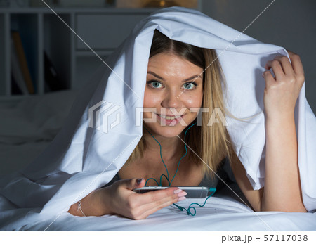 Female under blanket with mobile phone smiling on bed Female under blanket with mobile phone smiling on bed 57117038