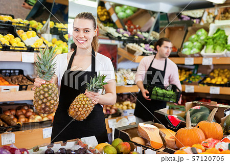 Positive saleswoman in apron offering fresh pineapples on the fruit store 57120706