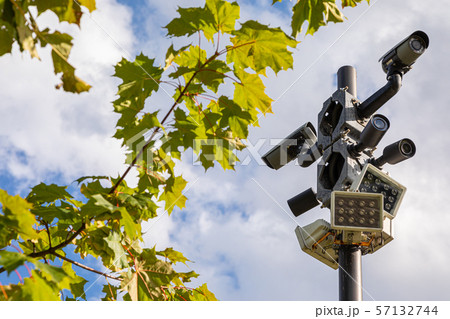 Black surveillance cameras and white street lamps on one column against sky and green leaves of Black surveillance cameras and white street lamps on one column against sky and green leaves of 57132744