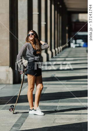 Beautiful young woman with longboard on the city street in sunny weather. Young hipster girl posing Beautiful young woman with longboard on the city street in sunny weather. Young hipster girl posing 57141744