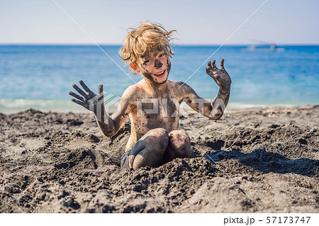 Black Friday concept. Smiling boy with dirty Black face sitting and playing on black sand sea beach Black Friday concept. Smiling boy with dirty Black face sitting and playing on black sand sea beach 57173747