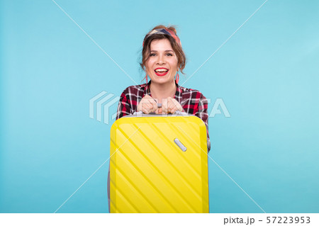 Positive young smiling woman in a plaid shirt holding a yellow suitcase posing on a blue background Positive young smiling woman in a plaid shirt holding a yellow suitcase posing on a blue background 57223953
