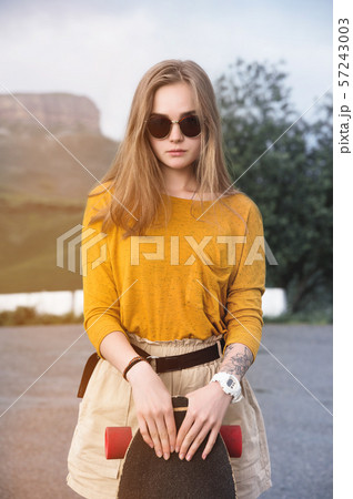 Beautiful and fashionable young woman in sunglasses and with a tattoo poses with a skateboard or 57243003
