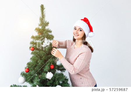 Holidays and celebration concept - Happy brunette woman in santa's hat decorates a christmas tree at Holidays and celebration concept - Happy brunette woman in santa's hat decorates a christmas tree at 57259687