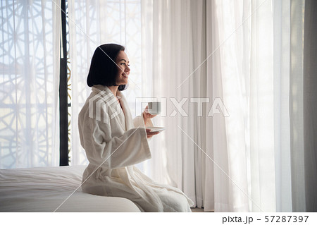 Attractive young girl in bathrobe holding a cup, looking at window and smiling while sitting on a 57287397