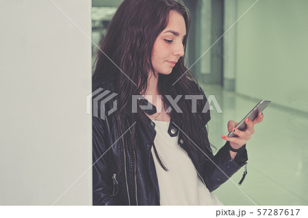 A beautiful girl uses a smartphone in an office building leaning against a wall A beautiful girl uses a smartphone in an office building leaning against a wall 57287617
