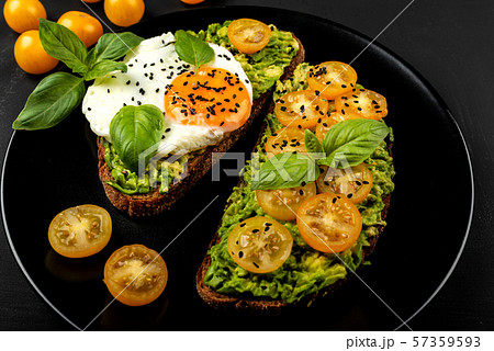 Open sandwiches with avocado guacamole, yellow cherry tomatoes, fried egg and basil on a black plate 57359593
