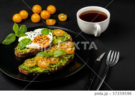 Open sandwiches with avocado guacamole, yellow cherry tomatoes, fried egg and basil on a black plate 57359596