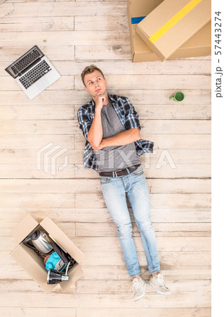 Young man lying on floor surrounded by boxes and laptop looking aside pensive top view Young man lying on floor surrounded by boxes and laptop looking aside pensive top view 57443274