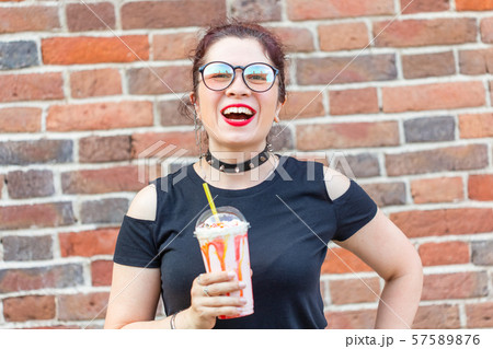 Young positive girl holds in Ruach a milkshake with a straw against a brick wall background. Summer Young positive girl holds in Ruach a milkshake with a straw against a brick wall background. Summer 57589876