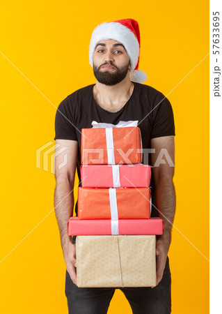 Disgruntled young man with a beard in a Santa Claus hat holds five gift boxes posing on a yellow 57633695