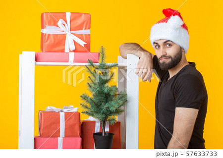 Young positive man with a beard and Santa Claus hat is standing near booth with gifts and Christmas 57633733