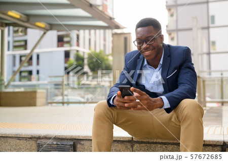 Young happy African businessman using phone while sitting outside the subway train station 57672685
