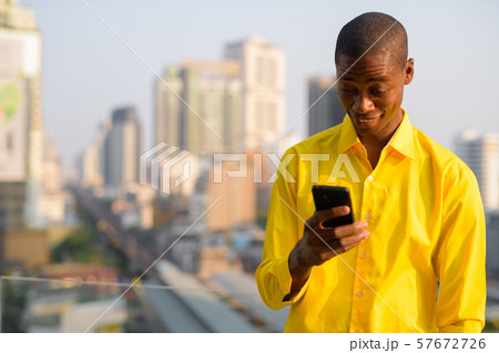 Happy young bald African businessman using phone against view of the city 57672726