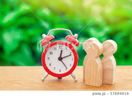 Three wooden human figures standing next to a red alarm clock on a nature background. Time concept. Business, Hourly payment. Man hours. Planning. minimalism 57694019