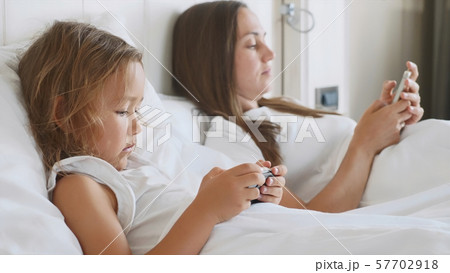 Young mother with little cute daughter are using gadgets lying on a white bed. Young mother with little cute daughter are using gadgets lying on a white bed. 57702918