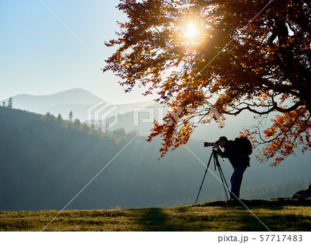 Hiker tourist man with camera on grassy valley on background of mountain landscape under big tree. Hiker tourist man with camera on grassy valley on background of mountain landscape under big tree. 57717483