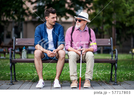 Young man and blind senior with white cane sitting on bench in park in city. Young man and blind senior with white cane sitting on bench in park in city. 57717803