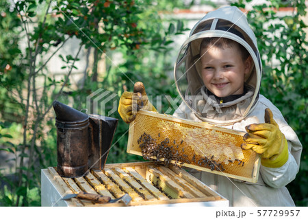 Cheerful boy beekeeper in protective suit near beehive. Honeycomb with honey. Organic food concept Cheerful boy beekeeper in protective suit near beehive. Honeycomb with honey. Organic food concept 57729957